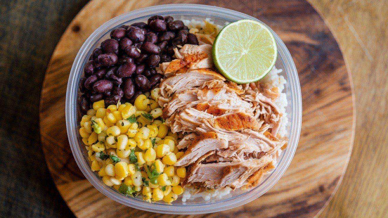 Six meal prep containers with colorful burrito bowls featuring shredded chicken, cilantro lime rice, black beans, corn, and fresh toppings