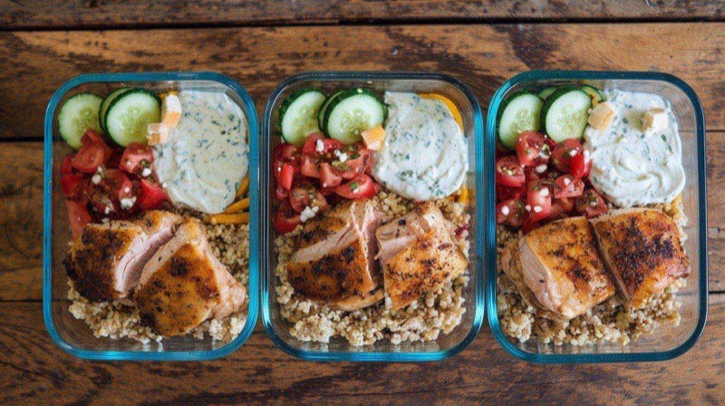 Five glass meal prep containers with Greek chicken bowls featuring golden chicken slices, fluffy quinoa, cucumber tomato salad, and white tzatziki sauce
