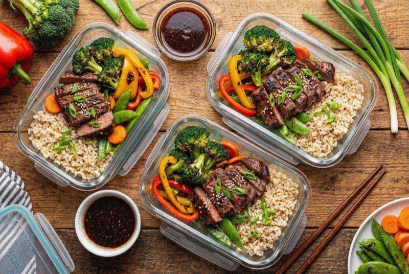 Meal prep bowl with sliced teriyaki-glazed steak, brown rice, grilled broccoli and zucchini, topped with sesame seeds and green onions