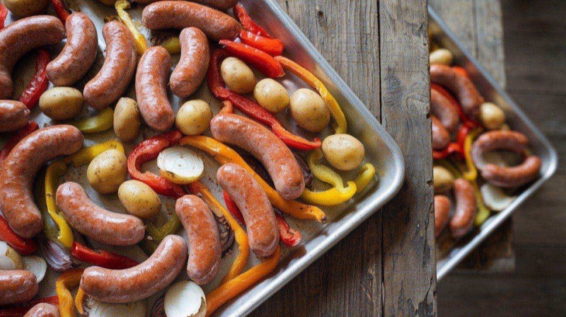 Sheet pan loaded with browned sausage rounds, colorful roasted bell peppers, red onion wedges, golden baby potatoes, and broccoli